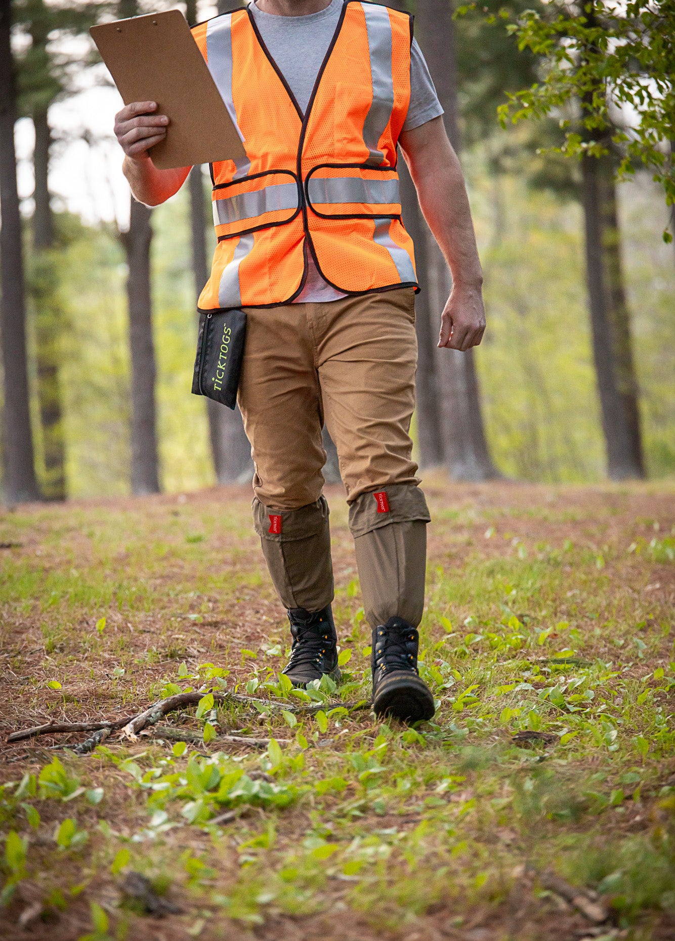 Surveyor with clipboard wearing safety vest, Ticktogs, hiking shoes, and Ticktogs storage bag clipped to belt loop.