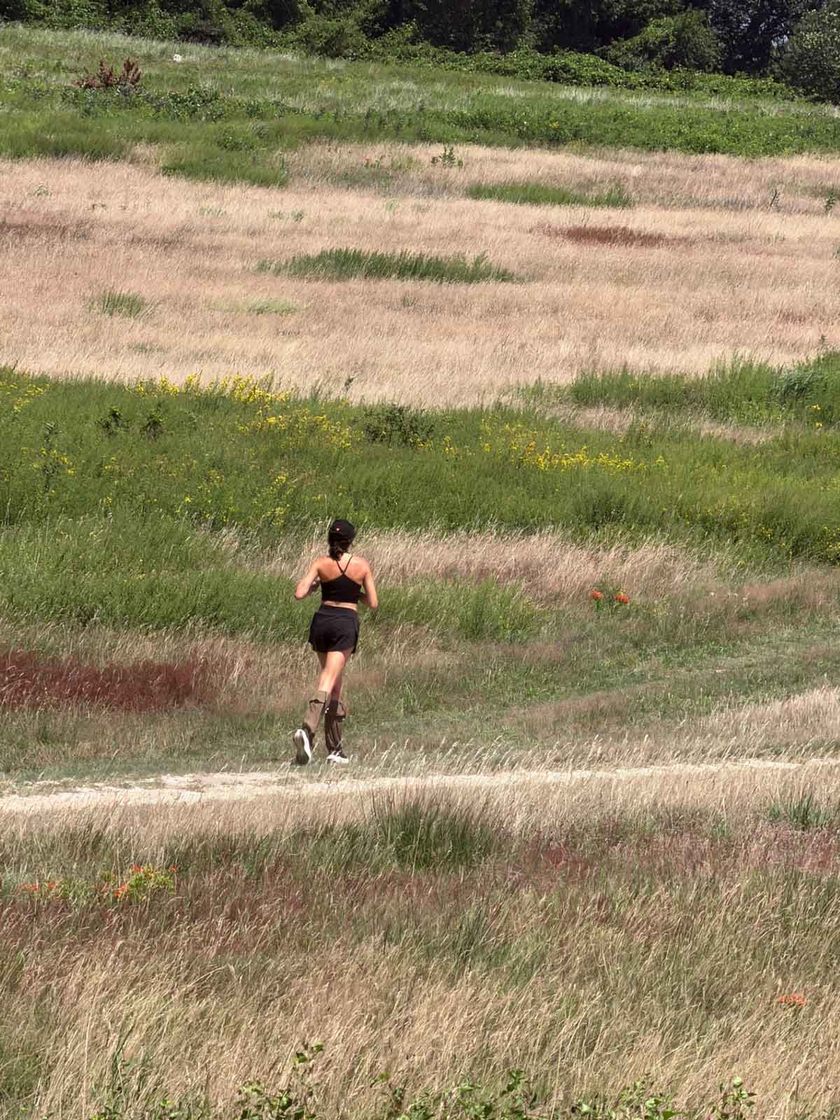 A woman running on a dirt trail through a grassy field, viewed from behind. She is wearing a black sports bra and black shorts and Ticktog oversocks