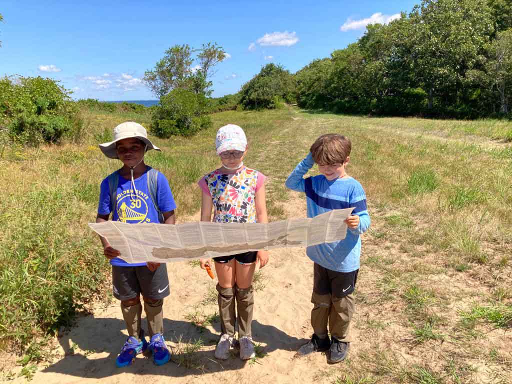 Three children standing outdoors on a dirt path holding up a large printed map in a grassy field with trees and blue sky in the background. The children are wearing Ticktog oversocks