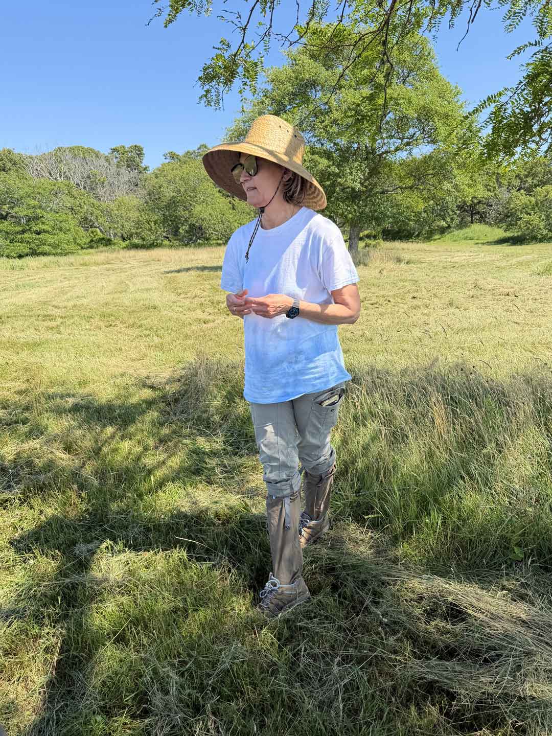 A person standing in a grassy outdoor field under trees, wearing a straw wide-brim hat, a light blue t-shirt, light gray rolled-up pants, and Ticktog oversocks.