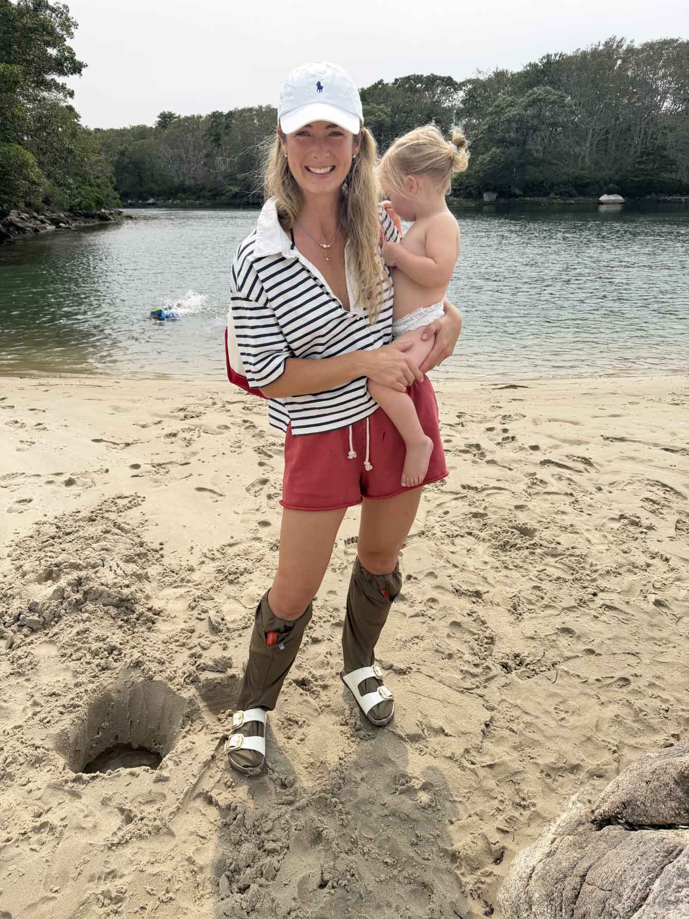 A woman standing on a sandy beach holding a toddler, wearing a white cap, a black and white striped long-sleeve top, red shorts, and Ticktog overshocks, with water and trees in the background.