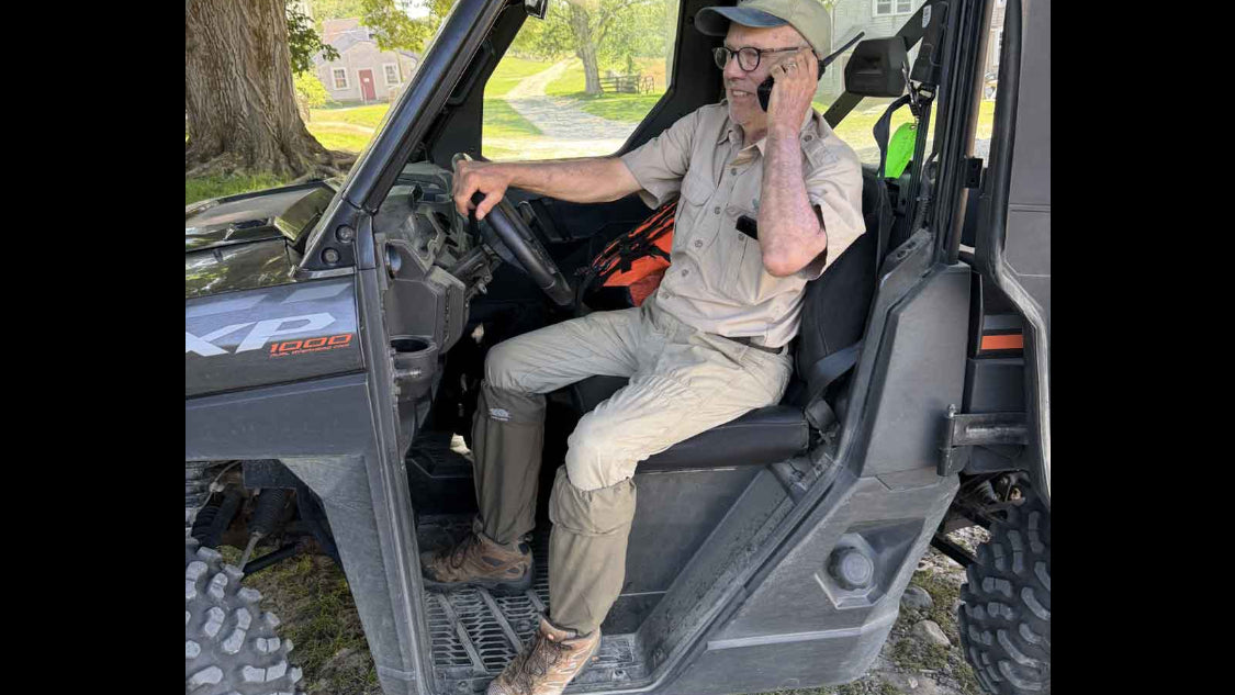 A man wearing a beige utility shirt and pants sits in the driver's seat of a black UTV, wearing ticktog oversocks, talking on a cell phone outdoors on a gravel driveway with trees in the background.