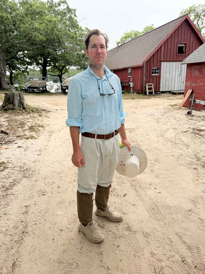A man standing on a sandy farm path in front of a red barn, wearing a light blue button-down shirt, cream pants, Ticktog oversocks, and holding a white hat.