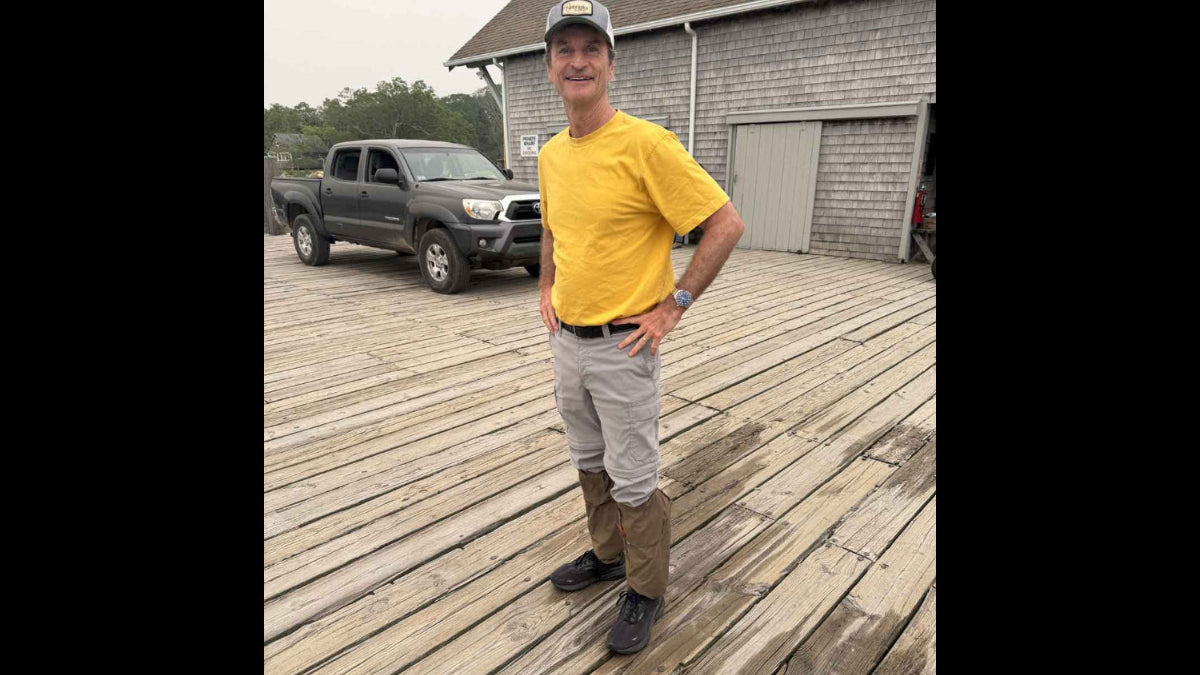 A man wearing a yellow t-shirt, light gray pants, and Ticktog oversocks stands on a wooden dock in front of a gray shingled building, with a dark pickup truck in the background.